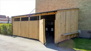 Bilden visar en tillbyggnad utav ett avfallsutrymme i trä. Avfallsrummet har en dörr för in- och utpassage.